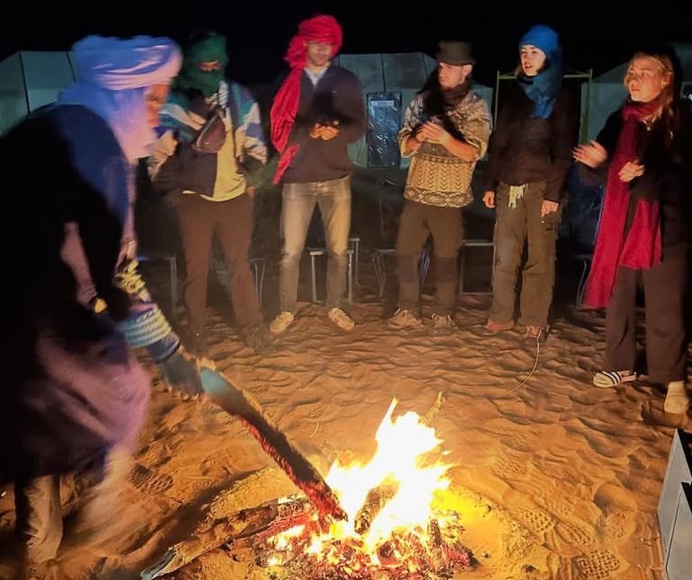 Night desert camp in Merzouga with travelers enjoying music and fire under the stars